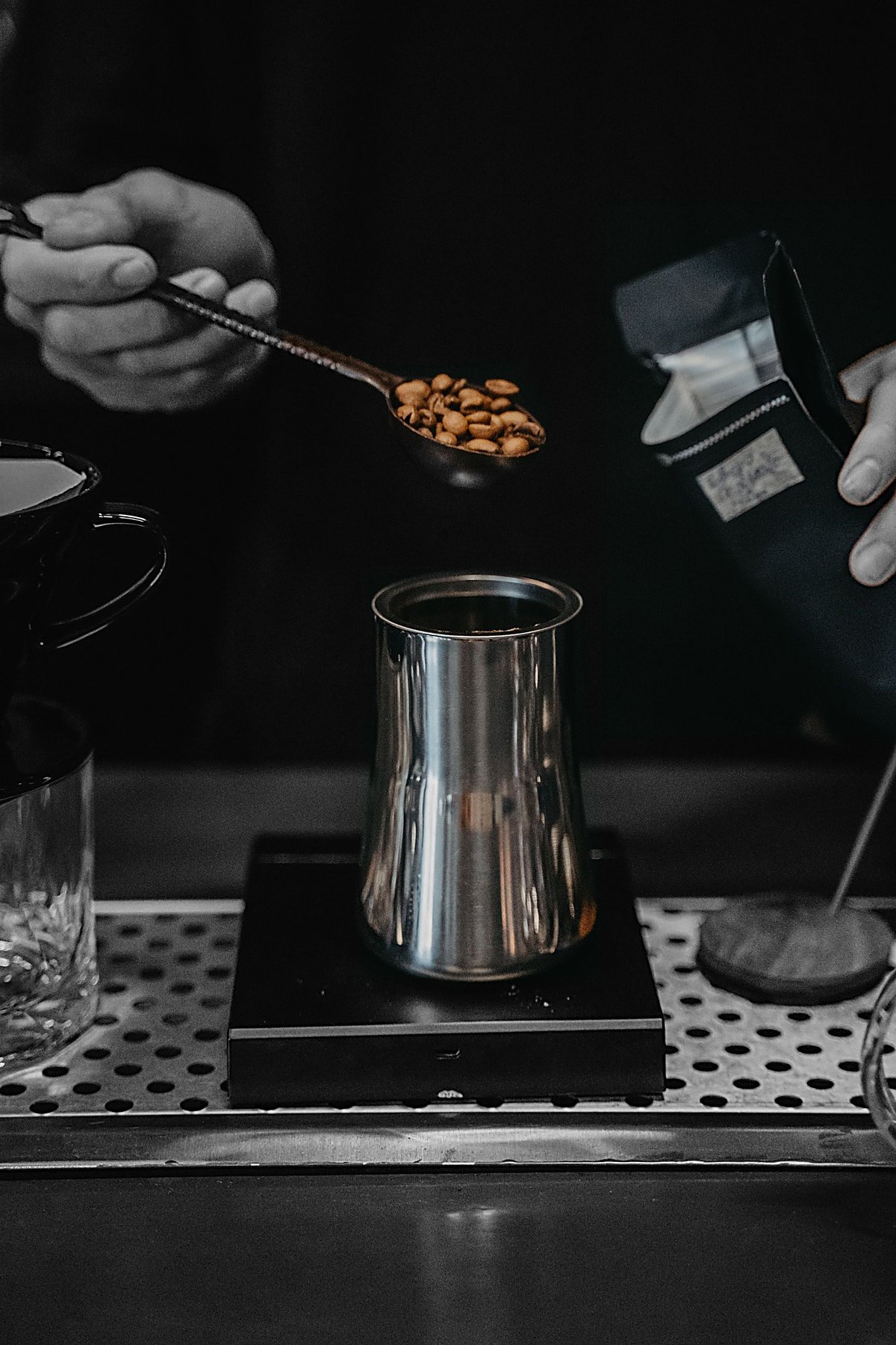 A barista scoops roasted coffee beans into a steel container on a scale, holding an open bag of coffee